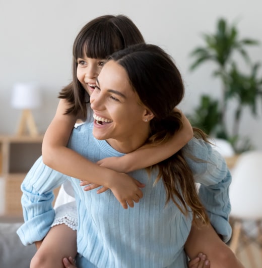 Mother Giving Piggy Back to Daughter in Their Living Room Enjoying Their Home Temperature From HVAC Services